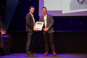 A full length shot of Jason Leslie and Chris Carter holding a certificate on stage looking straight at the camera