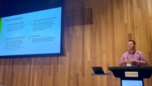 A photo of a man standing at a lectern which is in front of a large presentation screen