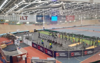 A photo of an indoor velodrome showing the track and a gym facility in the foreground